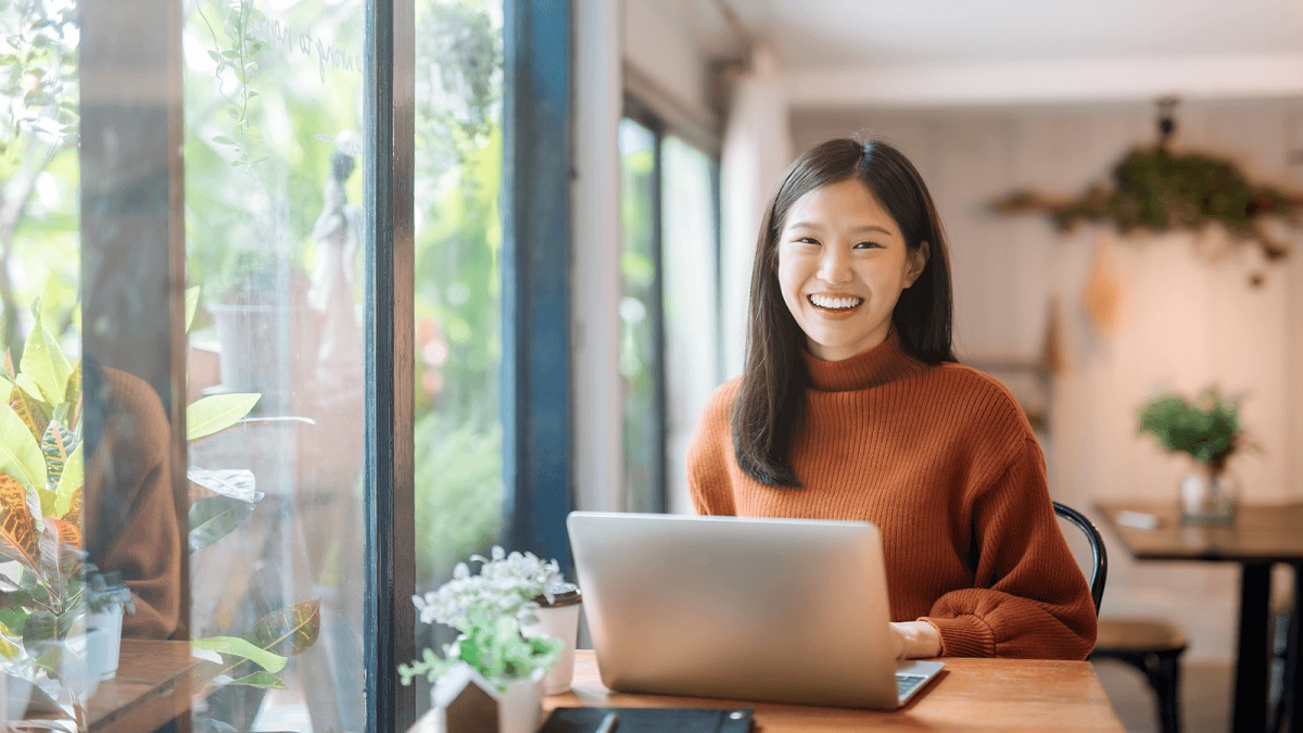 Empréstimo pessoal online em financeiras, mulher asiática com computador em uma sala cheio de vidros e arvores, com um semblante de felicidade
