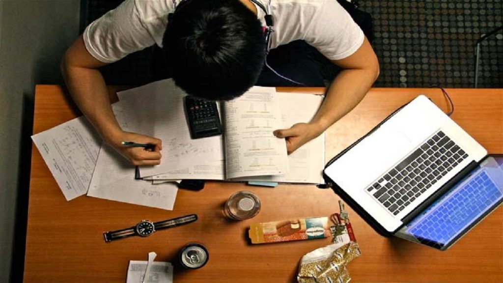 Uma foto vista por cima, no qual tem um homem estudando em uma mesa, a sua frente tem objetos para o estudo e ao lado um notebook. Estaudando para concursos públicos