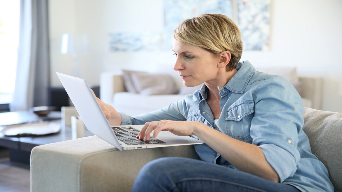 Mulher de camisa e calça jeans azul, sentada no sofá com o notebook olhando para a tela para ver as opções de empréstimo pessoal online em bancos.